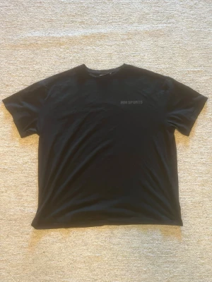 Svart t-shirt från MM Sports - Svart t-shirt från MM Sports med diskret logga framtill och stor logga i svart på ryggen. Klassisk rund hals och korta ärmar. Perfekt för dig som gillar en clean och sportig look.