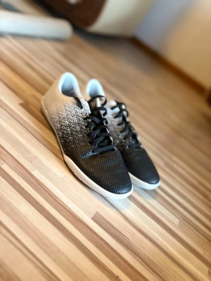 Svarta och vita fotbollsskor från SOC - Snygga fotbollsskor från SOC med en cool svart och vit gradientdesign. Skorna har svarta snören och en låg profil som passar perfekt för spel på inomhusplaner. Ovandelen är tillverkad i syntetmaterial för bra grepp och komfort.