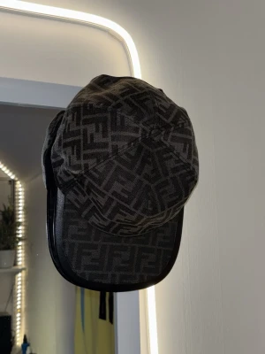 Svart Fendi keps med monogram - Säljer en svart keps från Fendi med det klassiska FF-monogrammet i grått över hela kepsen. Kepsen har böjd skärm och är tillverkad i ett tygmaterial som känns lyxigt och trendigt. Perfekt accessoar för dig som gillar exklusiva detaljer och streetstyle.
