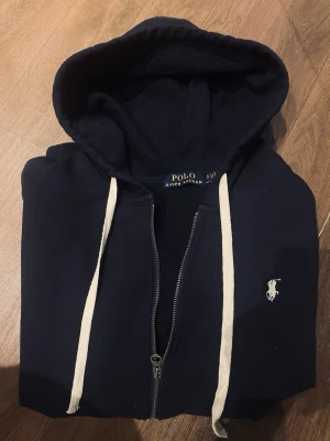 Marinblå hoodie från Ralph Lauren - Snygg marinblå hoodie från Ralph Lauren med vit logga på bröstet. Hoodien har dragkedja framtill, vit snörning i huvan och är tillverkad i mjukt material som känns riktigt skönt. Perfekt för dig som gillar klassisk och stilren design. Vid frågor kom PM