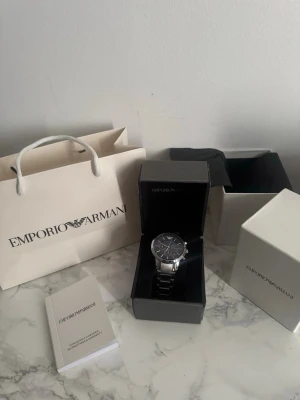 Emporio Armani silverfärgad klocka  - Stilren klocka från Emporio Armani med silverfärgad länk i metall och blått urtavla. Klockan har kronograf-funktion och tydliga indexmarkeringar. Levereras med originalask, påse och manual. Perfekt accessoar för en modern look. 