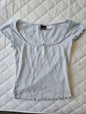Ljusblå ribbad topp från Gina Tricot - Supersöt ljusblå topp från Gina Tricot med ribbad struktur och liten rosett framtill. Toppen har korta ärmar med volangkant och en feminin, fyrkantig ringning. Perfekt för dig som gillar en enkel men söt stil. Använd max 2 ggr - mycket bra skick!