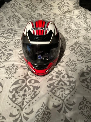 Takachi röd och vit motorcykelhjälm( Aldrig använd) Storlek L - Säljer en snygg motorcykelhjälm från Takachi i rött, vitt och svart Storlek L med coola grafiska mönster och stjärndetaljer. Hjälmen har visir och ventiler för bra luftflöde. Perfekt för dig som vill sticka ut på vägarna.