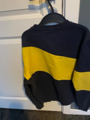 Mörkblå och gul sweatshirt från FILA - Cool sweatshirt från FILA i mörkblått med en bred gul diagonal rand över bröstet och armarna. Stor FILA-logga framtill i svart och rött. Tröjan har rund hals och ribbade muddar. Perfekt för en sportig och avslappnad stil.