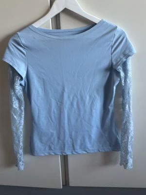 Ljusblå topp med spetsärmar - Trendig ljusblå topp med långa, transparenta spetsärmar. Toppens kropp är i mjuk bomull och har en enkel, rund halsringning. Perfekt för dig som vill ha en söt och stilren look med en twist av spets.