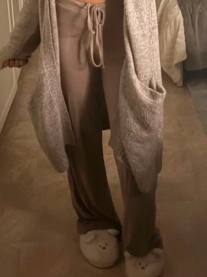 Beige loungewear-set med kofta - Mysigt loungewear-set i beige med vida byxor och matchande kofta med stora fickor. Byxorna har dragsko i midjan och koftan är stickad i mjukt material. Perfekt för chill hemma. Säljs utan tofflor.