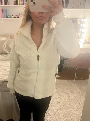 Vit teddyjacka med dragkedja - Mysig vit teddyjacka/tröja med hel dragkedja framtill och höga fickor med dragkedja. Jackan har en hög krage och är tillverkad i fluffigt teddy-material som ger en cool och avslappnad look. Perfekt för lager-på-lager. Mycket bra skick! Från Strova i strl M
