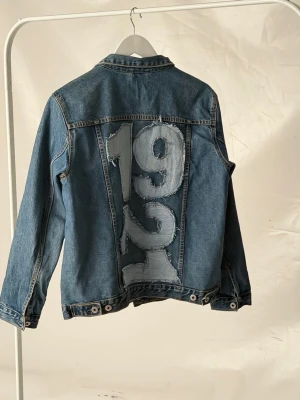 Jeansjacka med tryck - Jeansjacka i klassisk blå denim med stora siffror '1921' i lappad stil på ryggen. Perfekt för dig som vill sticka ut med en unik och trendig look.🩵