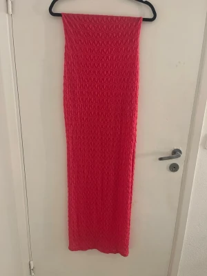 Rosa långklänning med strukturmönster - Trendig rosa långklänning med snyggt strukturmönster i tyget. Klänningen har en rak passform och är ärmlös, slits vid benet  vilket ger en stilren och modern look. Perfekt för dig som vill sticka ut med färg och enkel design. 