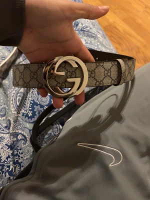 Gucci GG monogram bälte beige - Säljer ett klassiskt Gucci bälte med GG-monogram i beige och brunt. Bältet har ett stort, rundat GG-spänne i silverfärg och är tillverkat i skinn med canvasöverdrag. Perfekt accessoar för att lyfta vilken outfit som helst.