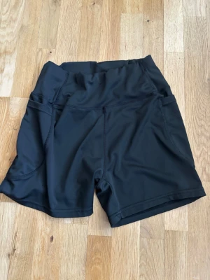 Svarta träningsshorts med hög midja - Snygga svarta träningsshorts med praktiska sidofickor. Tillverkade i stretchigt material med 90% polyester och 10% elastan för maximal komfort och rörelsefrihet. Perfekta för gymmet eller löprundan.