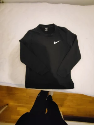 Svart Nike sweatshirt med logga - Svart sweatshirt från Nike med klassisk vit swoosh-logga på bröstet. Tröjan har rund halsringning, långa ärmar och ribbade muddar. Perfekt för en sportig och avslappnad stil.