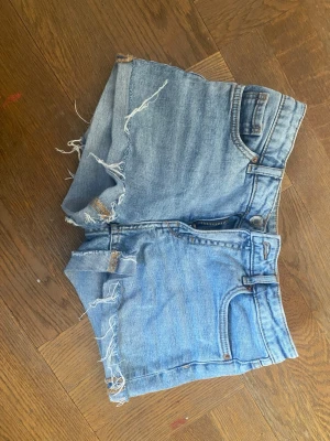 Ljusblå jeansshorts från Kappahl - Säljer ett par ljusblå jeansshorts från Kappahl med råa, fransiga kanter och klassisk femficksdesign. Shortsen har hög midja och stängs med dragkedja och knapp. Perfekta för varma dagar och en avslappnad stil.Low waiste 