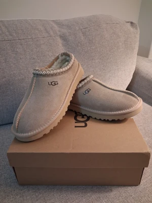 Beige fårskinnstofflor från UGG - Säljer ett par beige tofflor från UGG i mjukt mockaskinn med fårskinnsfoder. Klassisk slip-on modell med dekorativ kant runt öppningen och UGG-logga på sidan. 