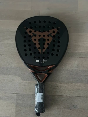 Wilson cupra Bela V2 padelracket svart/koppar - Säljer ett Wilson Bela padelracket i svart med kopparfärgade detaljer och signatur på bladet. Racketen har rund form, perforerat blad och grepp med plastskydd kvar. Snygg design och tydlig logga på sidan.