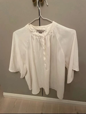 Vit lös blus med kort ärm - Snygg vit blus med avslappnad passform och korta ärmar. Blusen har en liten ståkrage och dekorativ knäppning framtill. Perfekt för en stilren och enkel look.