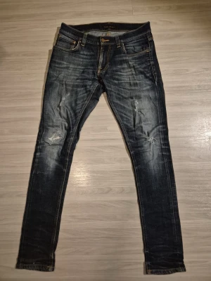 Nudie Jeans blå slitna jeans - Säljer ett par blå jeans från Nudie Jeans med snygga slitningar och tvättade detaljer. Modellen har smal passform och klassisk femficksdesign. Perfekta för dig som gillar en avslappnad men trendig stil. Modellen är Tight Terry, så skulle säga att den sitter som slim fit 