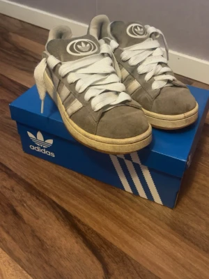 Adidas Campus grå sneakers i grå - Grå Adidas Campus sneakers i mjuk mocka med vita klassiska stripes och vit sula. Skorna har breda vita skosnören och Adidas logga på plösen. Snygg retrostil som passar till det mesta och ger en clean look.
