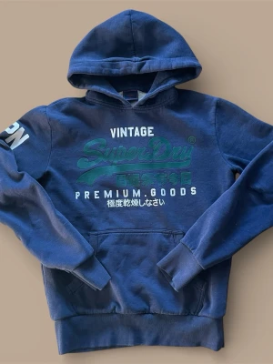 Mörkblå Superdry hoodie med tryck - Säljer en mörkblå hoodie från Superdry med grönt och vitt tryck på bröstet och ärmen. Tröjan har huva med snörning, känguruficka och ribbade muddar. Perfekt för chill dagar och streetstyle. Materialet är mjuk bomull och polyesterblandning. 