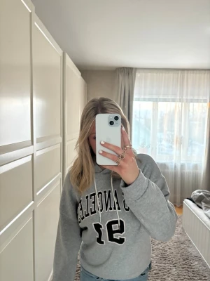 Grå hoodie med tryck 'Los Angeles 91' - Snygg grå hoodie med svart och vitt tryck där det står 'Los Angeles 91' på bröstet. Klassisk modell med huva och dragsnören, samt ribbade muddar vid ärmslut och midja. Perfekt för en avslappnad och trendig look.