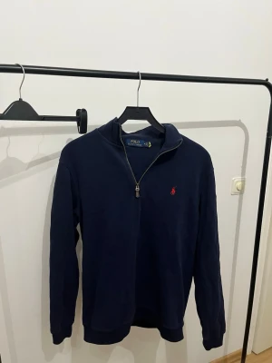 Mörkblå half-zip tröja Polo Ralph Lauren - Mörkblå långärmad tröja från Polo Ralph Lauren med half-zip och klassisk röd broderad logga på bröstet. Tröjan har hög krage och ribbade muddar vid ärmslut och nederkant. Perfekt för en stilren och avslappnad look.