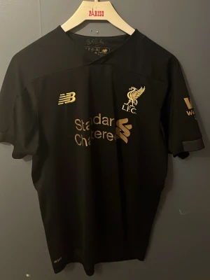 Svart Liverpool tröja New Balance - Snygg svart Liverpool FC tröja från New Balance med guldfärgade loggor och detaljer. Kortärmad modell med klubbmärke, sponsortryck och New Balance-logga på bröstet. Perfekt för dig som älskar fotboll och vill representera LFC. Tillverkad i lätt och funktionellt material.