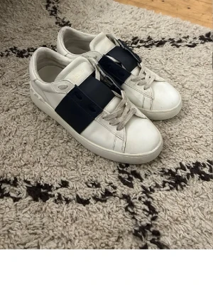 Valentino open - Säljer ett par vita Valentino Garavani open sneakers med bred marinblå läderdetalj över snörningen. Skorna har rund tå, platt sula och klassiska snören. Tillverkade i skinn och har diskret Valentino-logga på plösen. Perfekt för dig som vill ha en clean men lyxig look.
