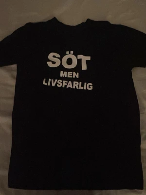 Svart t-shirt med texttryck - Cool svart t-shirt med vit text 'SÖT MEN LIVSFARLIG' på bröstet. Klassisk passform och rund halsringning, perfekt för dig som vill sticka ut med ett statement-plagg. 