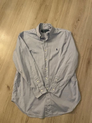Ljusblå skjorta från Ralph Lauren - Klassisk ljusblå skjorta från Ralph Lauren med button-down krage och broderad logga på bröstet. Skjortan har lång ärm, knappar framtill och är tillverkad i mjuk bomull. Skjortan är inte använd mycket, ca 3 gånger. 