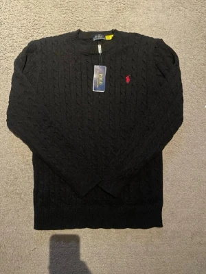 Svart kabelstickad tröja Polo Ralph Lauren - Kabelstickad svart tröja från Polo Ralph Lauren med rund hals och den klassiska röda logotypen broderad på bröstet. Tröjan har ribbade muddar vid ärmslut och nederkant. Perfekt för dig som gillar stilrena och tidlösa plagg.