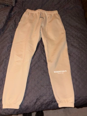Beige Essentials mjukisbyxor - Säljer ett par beige mjukisbyxor från Fear of God Essentials med logga tryckt på benet. Byxorna har resår i midjan och vid bensluten samt dragsko för justerbar passform. Perfekta för en avslappnad och trendig look.