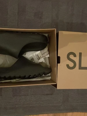 Svarta Adidas Yeezy Slide tofflor - Säljer ett par svarta Adidas Yeezy Slide tofflor. De har en chunky och modern design med räfflad sula och är tillverkade i ett mjukt, skönt syntetmaterial. Perfekta för en avslappnad och trendig look. ( använda en gång) om ni har några frågor är de bara att skriva privat
