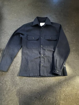 Jack n Jones Overshirt - Stilren mörkblå overshirt från Jack & Jones med klassisk krage, två bröstfickor med lock och knappar framtill. Tillverkad i ett kraftigare tyg som ger en snygg och avslappnad look. Perfekt att bära över en t-shirt eller hoodie.