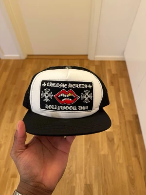 CH keps svart/vit - Svart och vit keps från med broderad patch framtill. Patchen har motiv av röda läppar, vita kors och texten ' Hollywood, USA'. Kepsen har mesh på sidorna och justerbar snapback.