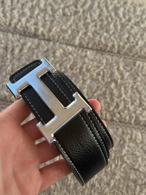 Svart H bälte  - Snyggt svart skärp med ett stort H-format spänne i silverfärgad metall. Skärpet är gjort av äkta skinn med vit kontrastsöm längs kanterna. Helt ny och är inte använd! Skicka till mig om det är någon fråga och skicka gärna bud om inte priset passar!!