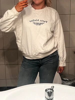 Vit sweatshirt Paris Atelier - Vit oversized sweatshirt med tryck 'Paris Atelier' framtill. Rund halsringning och ribbade muddar vid ärmar och nederkant. Perfekt för en avslappnad och trendig look.