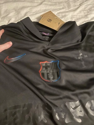 Svart FC Barcelona tröja Nike - Snygg svart FC Barcelona tröja från Nike med klubbmärke och Nike-logga i rött och blått på bröstet. Tillverkad i ett lätt och funktionellt material som passar perfekt till träning eller match. Modern design med diskreta mönsterdetaljer.