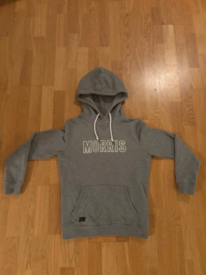 Grå hoodie från Morris med tryck - Snygg grå hoodie från Morris med vit logotyp framtill. Klassisk modell med huva och vita dragsnören samt magficka. Tillverkad i mjukt sweatshirtmaterial som är perfekt för chill dagar. Ribbstickade muddar vid ärmslut och nederkant. Inga defekter! Strl M passar S. Tveka inte att höra av er vid minsta fundering😊 pris ej hugget i sten.