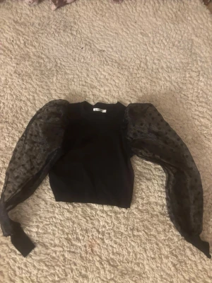 Svart topp med puffärmar och mesh - Trendig svart topp med croppad passform och stora puffärmar i transparent mesh med prickigt mönster. Perfekt statement-plagg för dig som vill sticka ut. Rund halsringning och åtsittande midja.