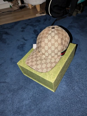 Beige Gucci keps - Säljer en beige keps från Gucci med det klassiska GG-monogrammet över hela kepsen. Kepsen har justerbart band bak och en detalj med röd och grön rand på sidan. Nästan aldrig använd och har inga fläckar, typ helt ny. Har också kvar lådan och två lappar som låg i när jag fick den.