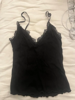 Svart spetslinne med tunna axelband - Säljer ett svart linne med tunna axelband och v-ringning. Linnet har fina spetsdetaljer längs urringningen och är tillverkat i ett mjukt, ribbat tyg. Perfekt att styla lager på lager eller bära som det är. Dock lite nopprig