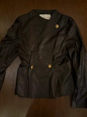 Svart skinnjacka från by Malene Birger - Snygg svart skinnjacka/ blazer med unik asymmetrisk knäppning och tre guldknappar framtill. Jackan har figurnära passform och långa ärmar, perfekt för dig som vill sticka ut med en stilren look.