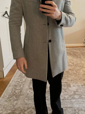 Ljusgrå ullkappa från J.Lindeberg - Stilren ljusgrå kappa från J.Lindeberg i ullblandning. Klassisk rak passform med knäppning framtill, diskreta fickor och en bröstficka. Perfekt för dig som vill ha en clean och modern look till hösten och vintern. Storlek 48/Medium