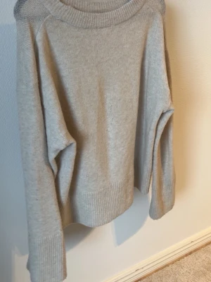 Beige oversized stickad tröja - Mysig beige stickad tröja med bred ribbad mudd vid ärmslut och nederkant. Tröjan har en avslappnad, oversized passform och rund halsringning. Perfekt för lager på lager och enkel att matcha med olika stilar. litet hål.