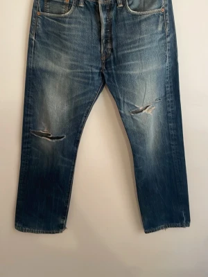  Levi's jeans 501 - Säljer ett par klassiska blå Levi's jeans med raka ben och tydliga slitningar på knäna och vid benslut. Jeansen har fem fickor, knappgylf och är tillverkade i kraftig denim. Perfekta för en avslappnad och trendig look.
