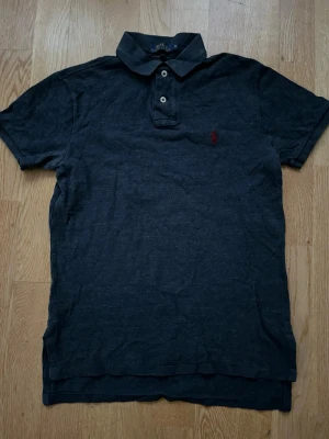 Mörkblå pikétröja från Ralph Lauren - Klassisk mörkblå pikétröja från Ralph Lauren med liten röd logga broderad på bröstet. Tröjan har korta ärmar, krage och knappar framtill. Tillverkad i mjuk bomull och har en snygg, enkel design som passar till det mesta.