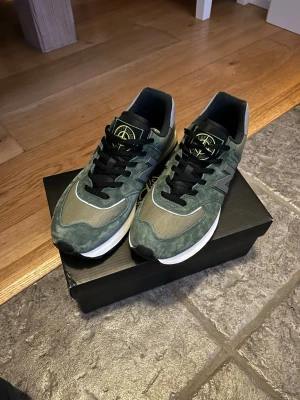Gröna Stone Island x New Balance sneakers - Snygga gröna sneakers från Stone Island x New Balance med svart och gul logga på plösen. Skorna har svarta skosnören, grå detaljer och vit sula. Ovandelen är i mocka och mesh för en sportig look. Perfekta för dig som gillar streetwear och unika samarbeten. Använda 1 gång