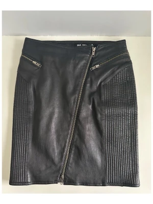 Svart skinnkjol med dragkedjor BLK DNM - Säljer en svart kort skinnkjol från BLK DNM i storlek 36 med sned dragkedja framtill och två dekorativa dragkedjor upptill. Kjolen har ribbade detaljer på sidorna som ger en cool biker-look. Högkvalitativt veganskt konstläder. Perfekt för dig som vill ha en edgy stil. Mycket gott skick!