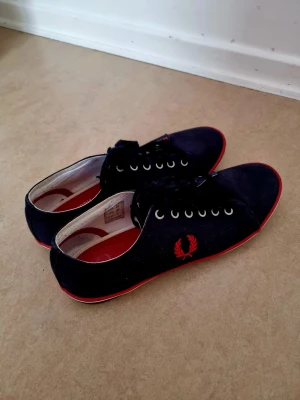Fred Perry sneakers stl43 - Ett par Fred Perry sneakers i blå färg med röda detaljer. Skorna har snörning och en klassisk design. Perfekt för en avslappnad och stilren look. Storlek 43