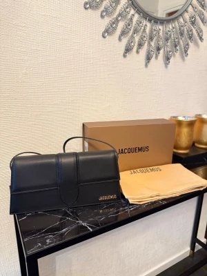 Jacquemus Le grand Bambino läderväska - Stilren svart kuvertväska från Jacquemus i slätt läder med guldfärgad logga på framsidan. Väskan har ett elegant lock och ett smalt handtag upptill. Perfekt för dig som gillar minimalistisk och trendig design.